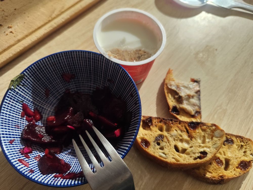 Un reste de betteraves rouges dans un petit bol bleu avec une fourchette au-dessus dessus. Des tranches de pain gratinées et un pot de rillettes devant une planche de bois sur un plan de travail de cuisine.