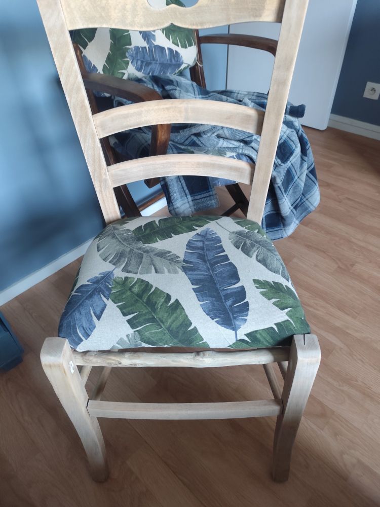 Une chaise en bois brut pâle dont l'assise est composée d'un tissu écru et de feuilles de bananier vertes et bleues devant une autre chaise sur laquelle est une robe de chambre à carreaux. Le sol est du lino imitation bois.
