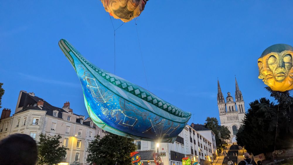 Un ballon en forme de drakkar bleu survole la foule sur fond de ciel bleu un soir au pied de la cathédrale d'Angers lors des derniers " Accroches coeurs".