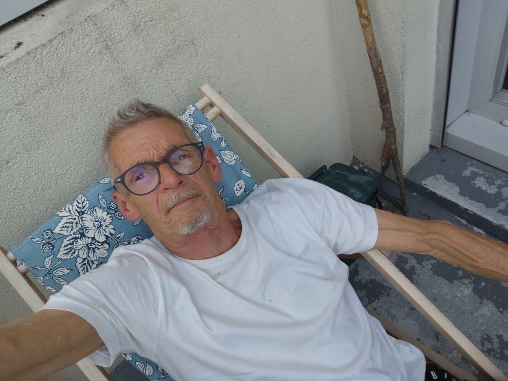 Homme mûr, cheveux gris, lunettes en t-shirt  blanc sur une chaise longue toile bleue à fleurs blanches, se prend en selfie 