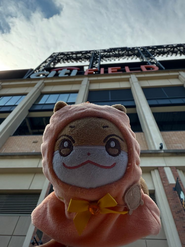 A sinister looking Jjoongrami in a cape in the foreground. The camera is aimed upward at the Citi Field sign on the stadium. A partly cloudy sky is above that. 