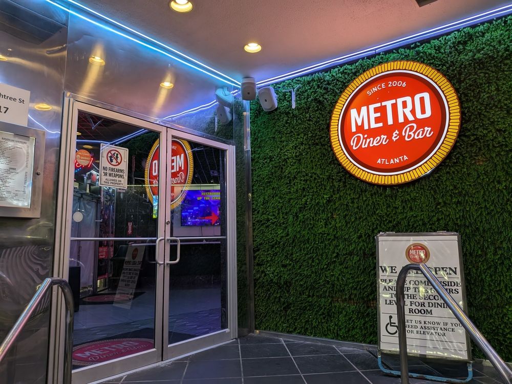 The entrance to "METRO Diner & Bar" is shown. On the right, a large, circular, orange and red sign with "METRO Diner & Bar ATLANTA" in white text hangs against a vibrant green artificial foliage wall. The text "SINCE 2006" is at the top of the sign. To the left, a glass double door entrance with horizontal push bars is visible. Above the doors, bright blue neon lights illuminate a metallic ceiling. A sign on the door indicates "NO FIREARMS OR WEAPONS." A black doormat is on the floor in front of the doors, and metal railings lead down to the entrance. A white sign with red text for Metro Diner & Bar is also visible near the entrance.
