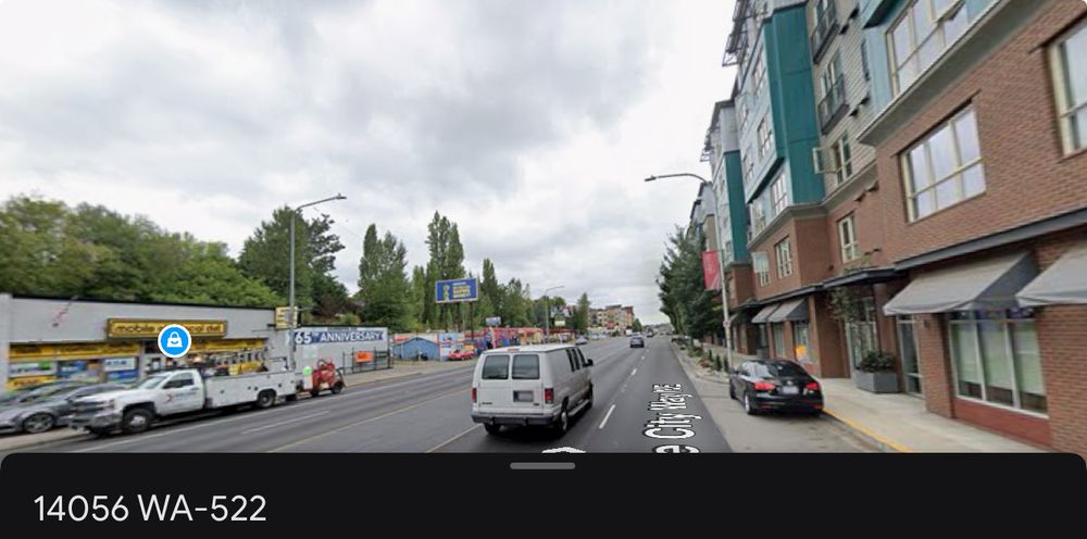 140th/143rd and Lake City Way looking south. 1 large 5 story apartment building is on the right. It spans about 2 blocks. The apartment goes right up to the narrow sidewalk and stroad. The view out the window of apartments on this side of these double loaded corridor buildings is solely Lake City Way. There are only a few medium sized street trees in view after about a full block, where for a full block there are none at all.