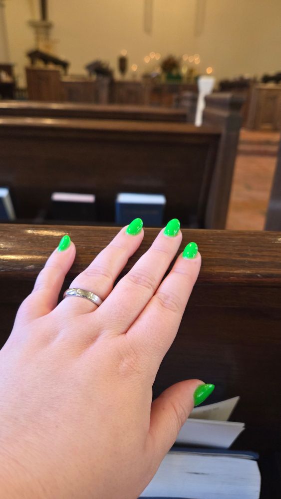 My hand on a wooden pew. I have a green manicure and a silver wedding ring. You can see the altar in my parish with lit candles blurry in the background and some pew books and the pews in front of me and a brick floor. 