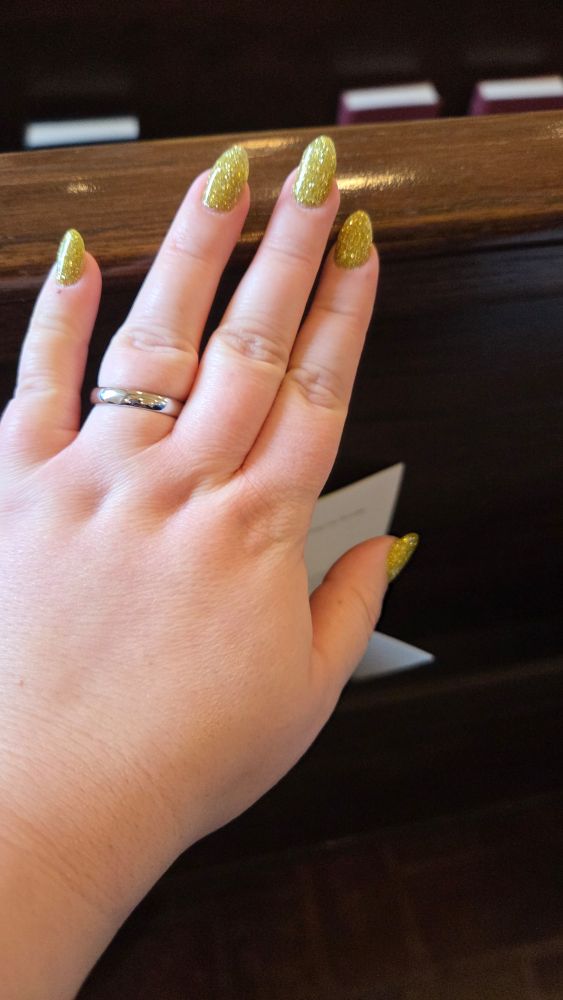My hand on a wooden pew. I am wearing a silver wedding ring and I have gold flitter nails. You can also see a program and pew books. 