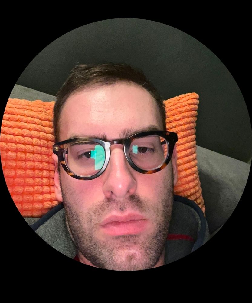 The current profile picture of article author Josh Greenblat, a white man, wearing glasses reflecting a screen and laying on some pillows. He is not smiling. 
