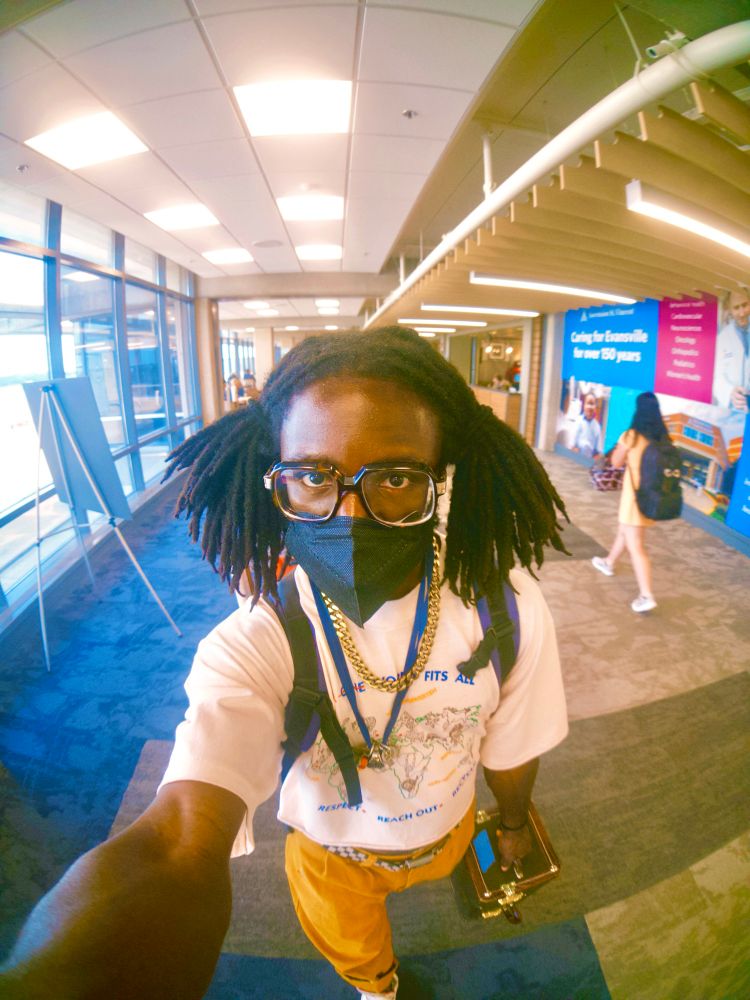 Selfie of JER walking through an airport. Their hair is in pigtails and they’re wearing a mask. There holding the phone in front of them with their right hand, a trumpet case is in their left 