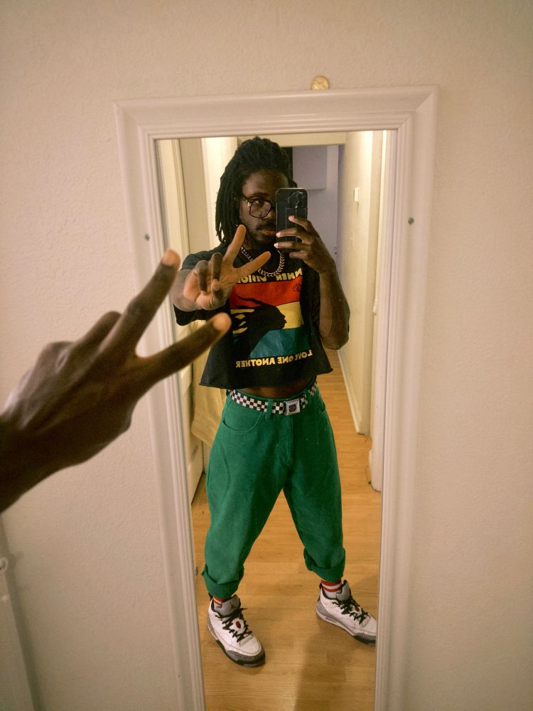 JER taking a full body selfie in the mirror. In the foreground is their hand throwing a peace sign, coming in from behind the camera. There’s a mirror or a white wall and in the reflection is JER holding he phone throwing the peace sign. They have white sneakers, green pants, a checkered belt, and a black crop top with a red yellow and green design. 