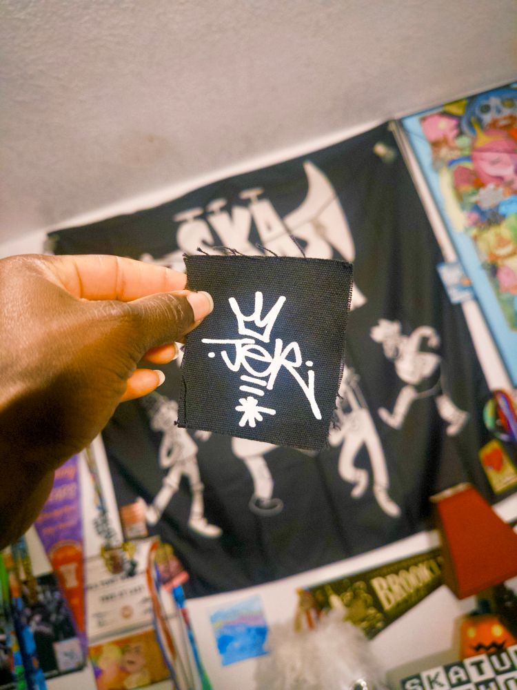 A picture of Jer’s hand holding up a JER patch. The patch is black cloth with a white print. The print is the JER tag logo