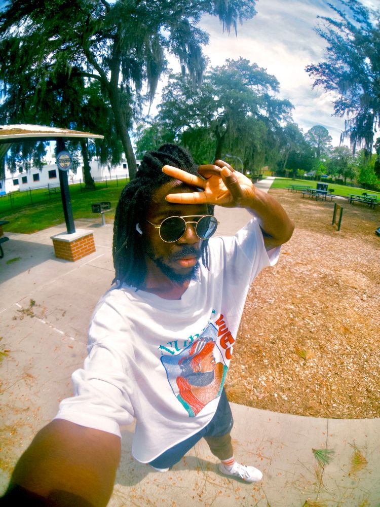 Selfie of JER walking and throwing a peace sign up in front of their forehead. A side walk, pavilion, building, and greenery fills the background 