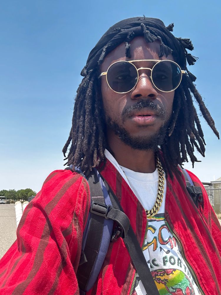 Selfie of JER wearing a white shirt with a red baseball jersey over it. Sunglasses, and a satin cap with their locs coming out of the sides 