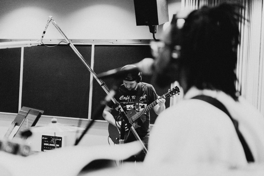 Black and white image of Ricky playing guitar. Microphone stands obscure part of Ricky as the camera focuses on him. In the foreground is an out of focus shot of her singing into a microphone. 