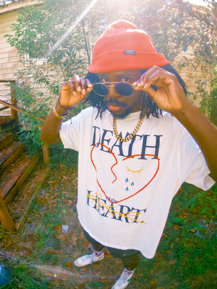 A picture of JER wearing the white death of the heart shirt, lowering their sunglasses to look at the camera 