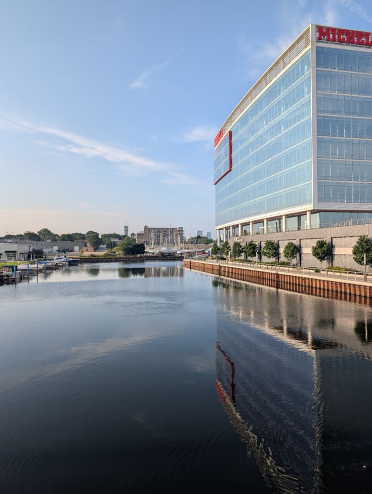 A picture of a river with buildings in the distance. The right side of the river is lined by trees and a big rectangular window covered building. The river reflects everything clearly