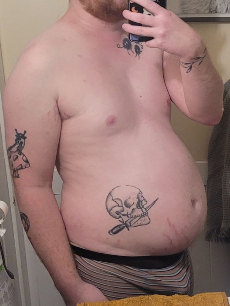 A young man standing shirtless with several tattoos. His belly bulges in front of him with stretch marks creeping up from underneath. 