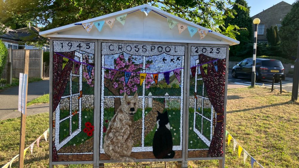 Welldressing. Large boards saying 1945-Crosspool-2025 and a depiction made of small elements of natural materials featuring a terrier type dog, head turned towards the viewer, and a black and white cat. They are at a window looking out at a gate and garden path, there is bunting.