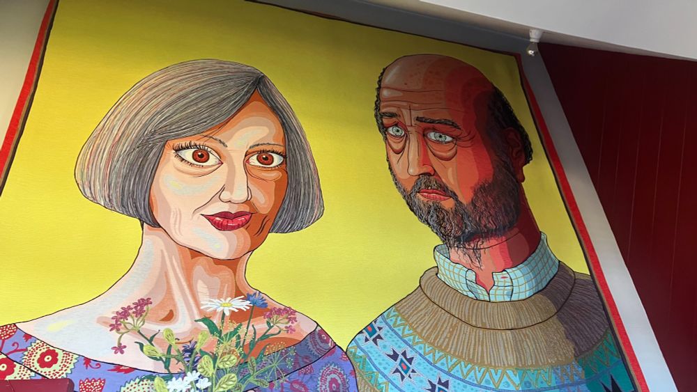 A middle-aged couple, a woman with grey bobbed hair and a balding bearded man in tapestry by Grayson Perry. Bright yellow background.