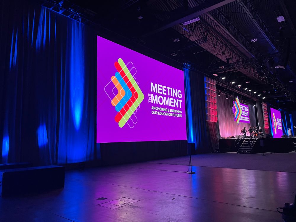 Blue curtains flank a purple projection that says “Meeting the Moment; anchoring and enriching our education futures” with a stage in the background on the right