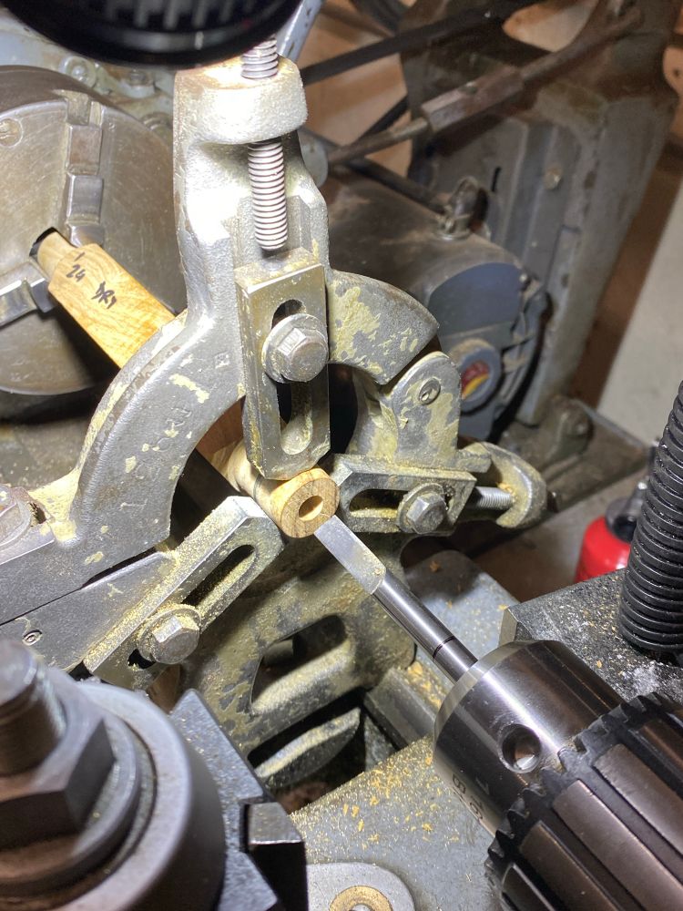 Piece of wood held between a chuck and a steady rest on a metal lathe. The piece is bored and right by the hole we can see the D-bit that made it. The bit is held in the Jacobs chuck in the tail stock. 