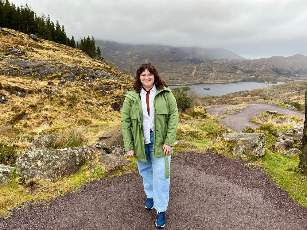 Erin stands in Killarney National Park with rocky hills, pine trees and a river in the distance behind her. She has brown, shoulder length hair with bangs and is wearing a white button down shirt, green raincoat and light blue jeans. 