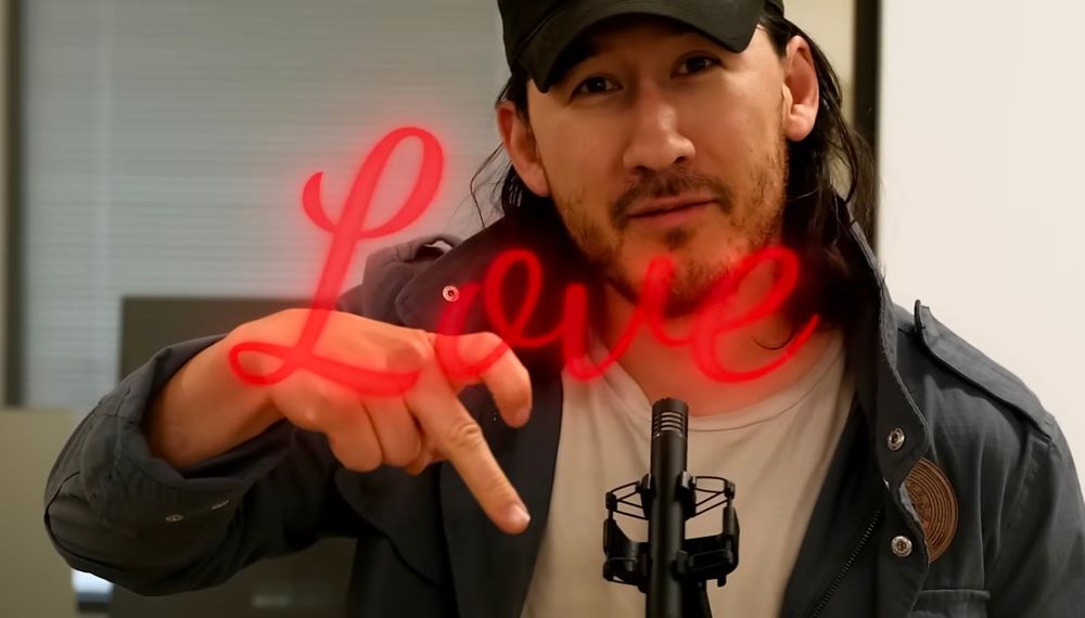 screenshot of youtuber Markiplier forming a half heart shape with his index and middle finger while staring into the camera. the word "Love" is displayed over the image in a red, cursive font.