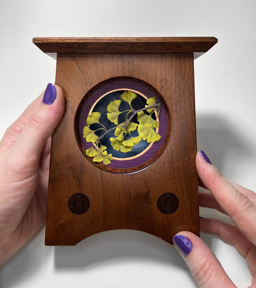 Cut paper ginkgo leaves framed in a wooden clock case, with purple and gold circular mats, and blue background. The piece is being held to show scale. 