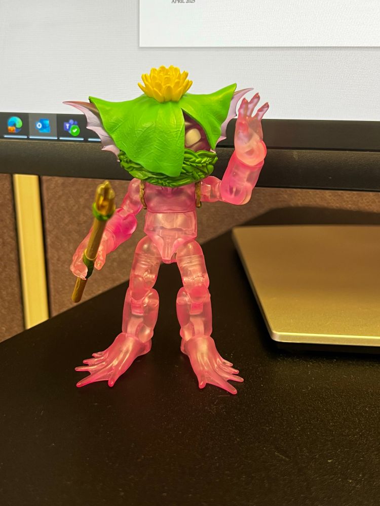 A small figurine “plunderling”. It appears to be a translucent pink goblin creature wearing a lily pad hat, a leafy face mask, and holding a spear. It’s waving at the camera! 