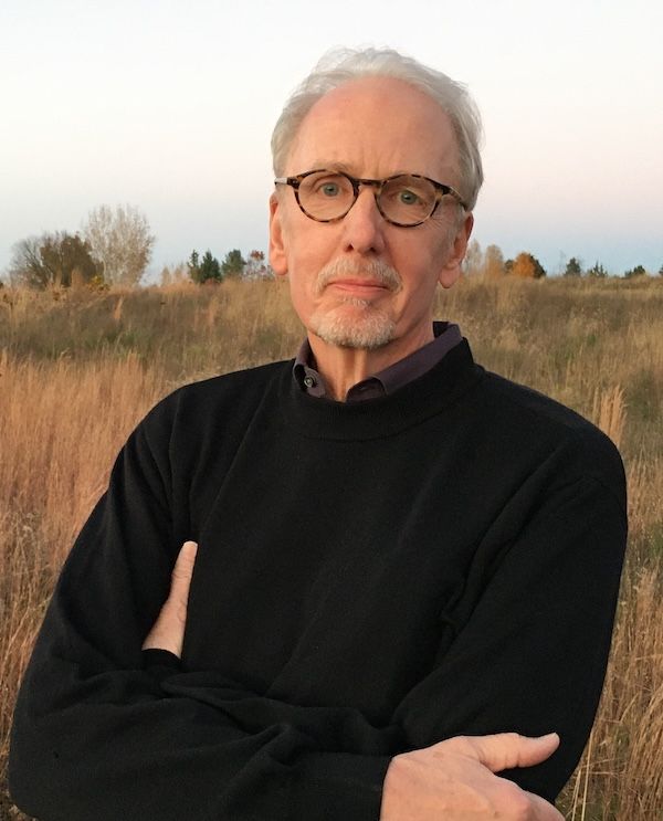 Photo of a man with white hair, a short-cropped beard and mustache, and glasses standing in a field. He is wearing a black sweater.