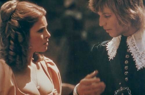 Raquel Welch as Constance and Michael York as D'Artagnan. They stand close together, her face etched in worry while his gaze lingers on her very prominent bosom. She is wearing a deep cut gown of peach silk, with puff sleeves, and her auburn hair is pulled half back into a bun while the rest hangs in voluminous ringlets. He wears a black silk doublet with jet inlaid metal buttons, a wide lace edged collar, and his blonde jaw-length hair is tousled and poofy. 