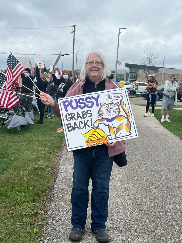 Woman holding 3 American flags and a sign reading “Pussy grabs back!” It depicts a calico cat with its claws on Trump’s head.