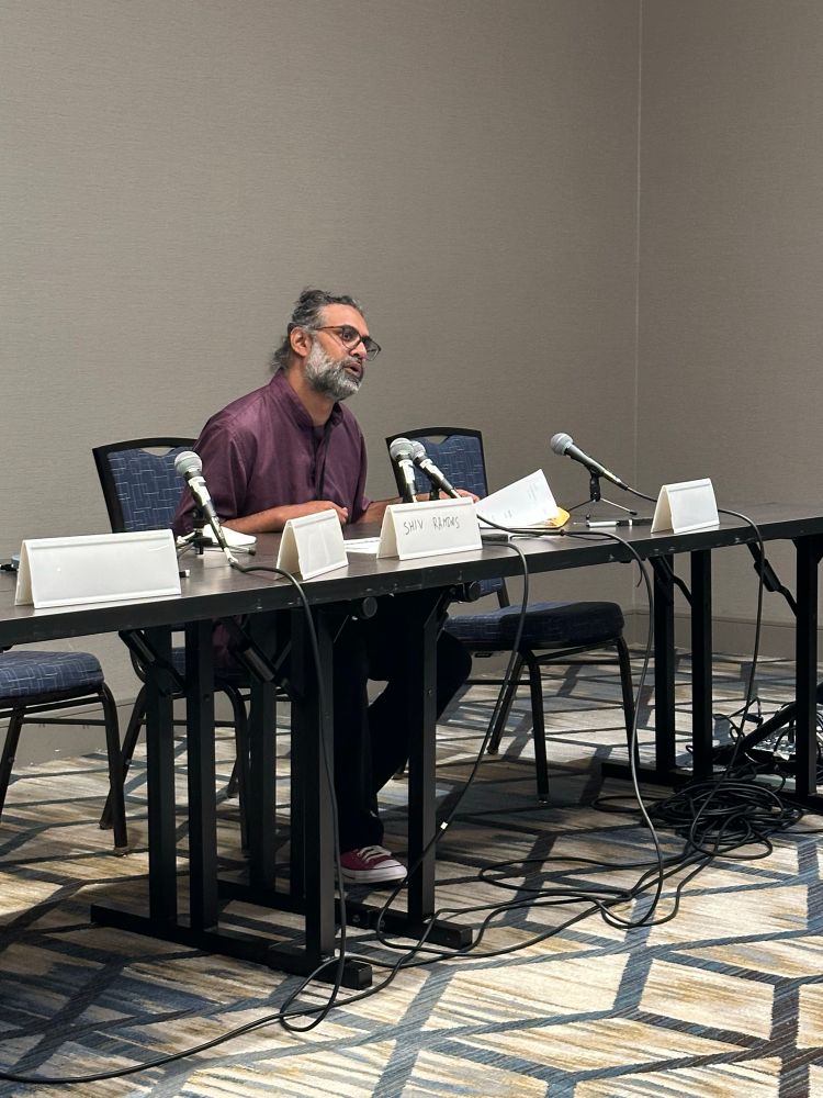 Shiv Ramdas in dark purple shirt reads into a microphone at the center of a wide table with multiple microphones.