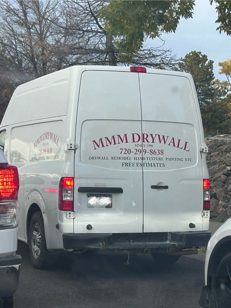 White utility van on the road, the rear window is advertising a business in large Red, All caps text that says: “MMM DRYWALL”. Subheading, also in red, says: “since 1990”. Text underneath, much smaller than the title says, “Drywall, Remodel, Hand Texture, Painting, Etc. FREE ESTIMATES”