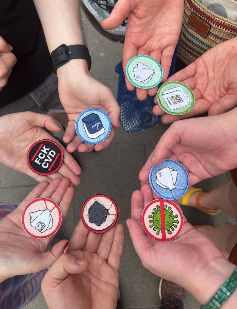 In einem Kreis halten acht Hände jeweils einen gestickten Patch. Alle Patches sind rund. Man sieht zwei Patches mit hellgrünem Hintergrund und grünem Ring drumrum, in der Mitte ist beim einen ein aranet4 CO2-Messgerät und beim anderen eine 3M Aura Maske mit weißen Kopfbändern zu sehen. Man sieht zwei Patches mit hellblauem Hintergrund und blauem Ring drumrum, in der Mitte ist beim einen ein Pluslife Testgerät und beim anderen eine 3M Aura Maske mit blauen Kopfbändern zu sehen. Man sieht zwei Patches mit hellrotem Hintergrund und rotem Ring drumrum, in der Mitte ist beim einen eine schwarze Trident Maske und beim anderen eine 3M Aura Maske mit Ventil und roten Kopfbändern zu sehen. Ein weiterer Patch hat einen roten Ring drumrum und einen schwarzen Hintergrund, auf dem zwischen zwei roten Balken oben und unten in der Mitte in weißer Farbe die Buchstaben FCK CVD stehen. Ein weiterer Patch zeigt auf hellrotem Hintergrund ein grünes Virus, was von einem roten Ring umgeben und durchgestrichen ist.
Man sieht im Hintergrund noch die Schuhe der Personen und den grauen Pflasterboden draußen vor einem Laden.