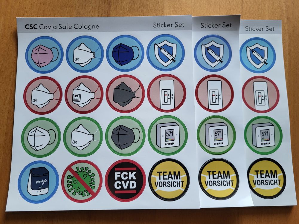 Zu sehen sind auf einem Tisch aufeinanderliegende Stickerbögen. Am Kopf des Bogens steht "CSC Covid Safe Cologne" und "Sticker Set". Es sind 16 unterschiedliche Sticker auf einem Bogen, 4 mal 4 Stück. In der ersten Reihe sind eine rosa FFP2 Maske, eine 3M Aura FFP2 Maske, eine dunkelblaue FFP2 Maske und ein Schutzschild mit einer Spritze zu sehen, alle auf hellblauem Hintergrund mit blauem Ring drumrum. In der zweiten Reihe sind eine 3M Aura FFP3 Maske, ohne und mit Ventil, eine schwarze Trident FFP3 Maske und ein negativer Schnelltest, alle auf hellrotem Hintergrund mit rotem Ring drumrum. In der dritten Reihe sind eine weiße FFP2 Maske, eine 3M Aura FFP2 Maske mit weißen Bändern, eine schwarze FFP2 Maske und ein aranet4 CO2-Messgerät, alle auf hellgrünem Hintergrund mit grünem Ring drumrum. In der vierten Reihe sind ein Pluslife Testgerät, bei dem das grüne Licht leuchtet, ein durchgestrichenes Virus, ein Schriftzug mit den Buchstaben FCK CVD in zwei Zeilen übereinander und ein Schriftzug "Team Vorsicht" mit einer Maske im Hintergrund.
