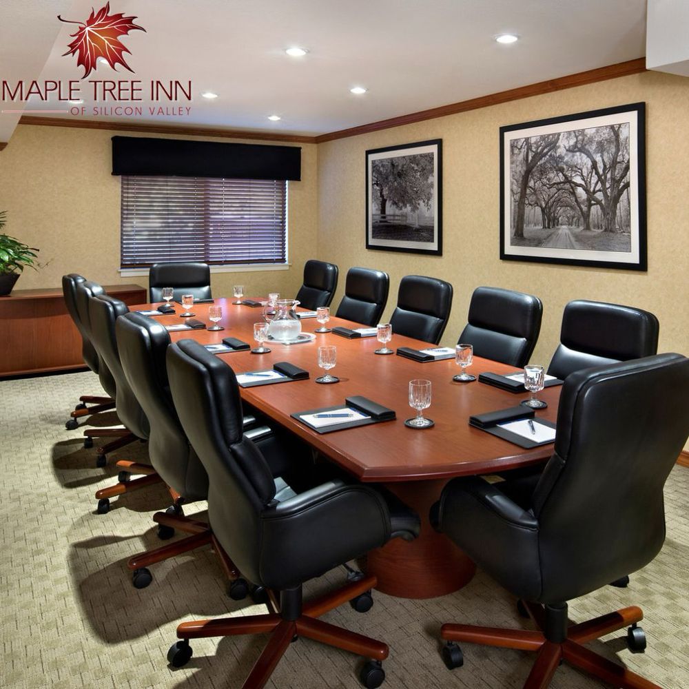 A carpeted room with 12 leather chairs around a long oak desk set with water glasses and paper with pens. Black and white photography adorns the wall with a window in the back. The logo that reads Maple Tree Inn of Silicon Valley is in the upper left corner with a picture of a maple leaf. 