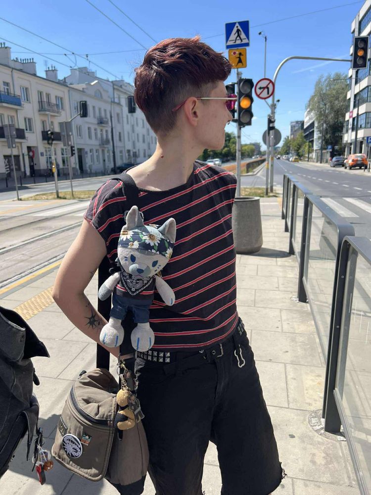 remi, a skinny white butch in shorts and a black and red striped top. babirousa's dog izzy is attatched to their bag strap with a bandana collar, and wearing blue pants and a tank top made from remi's shirt material. there's a german shepherd plush keychain on the bag, as well as ram ranch's ed and izzy keychains 