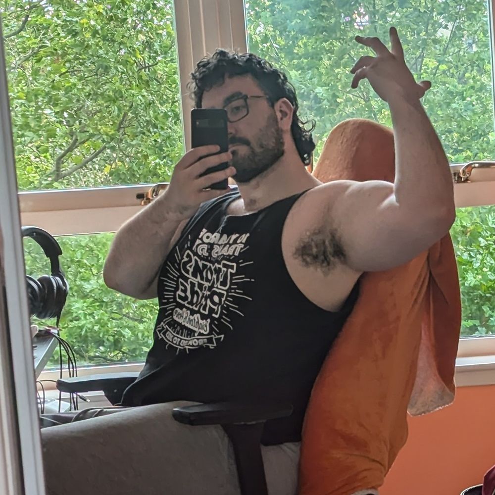 A large masc presenting person sits at a desk flexing a very impressive arm 