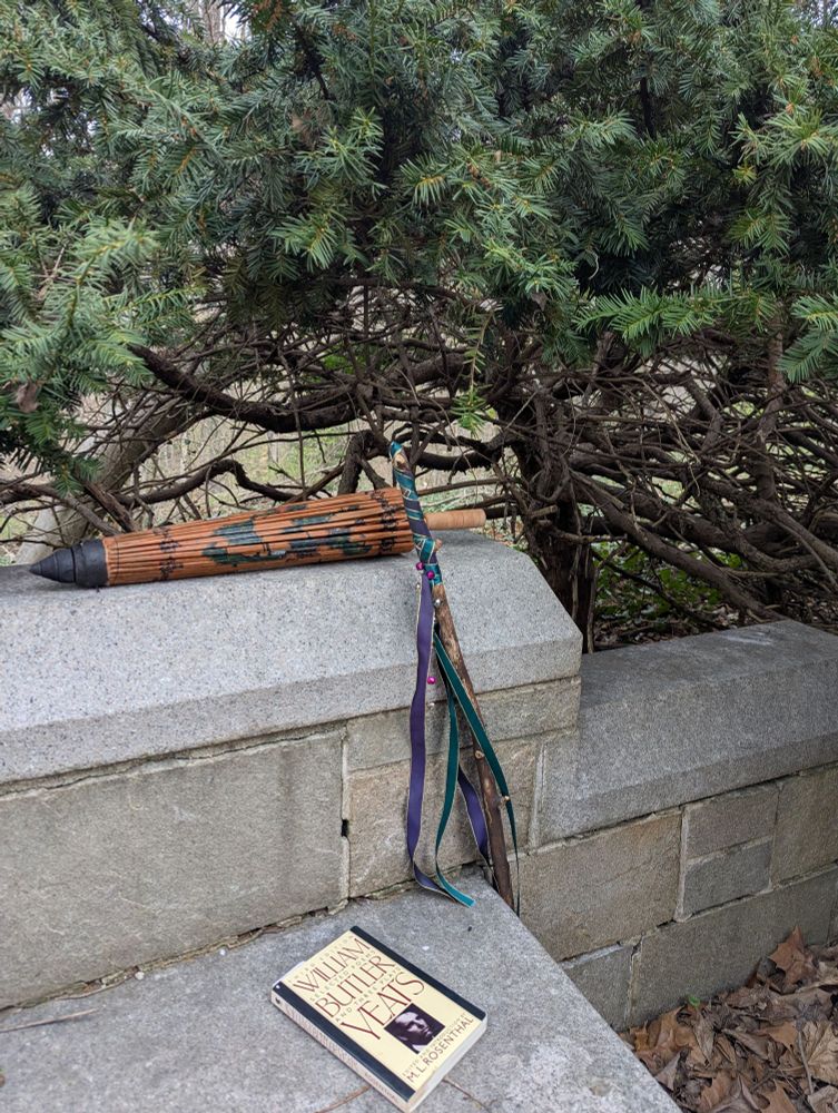 A volume of poetry by W.B. Yeats on a bench, a walking stick with purple and green ribbons leaning against the bench, and a bamboo sunshade on the wall, with a tree in the background.