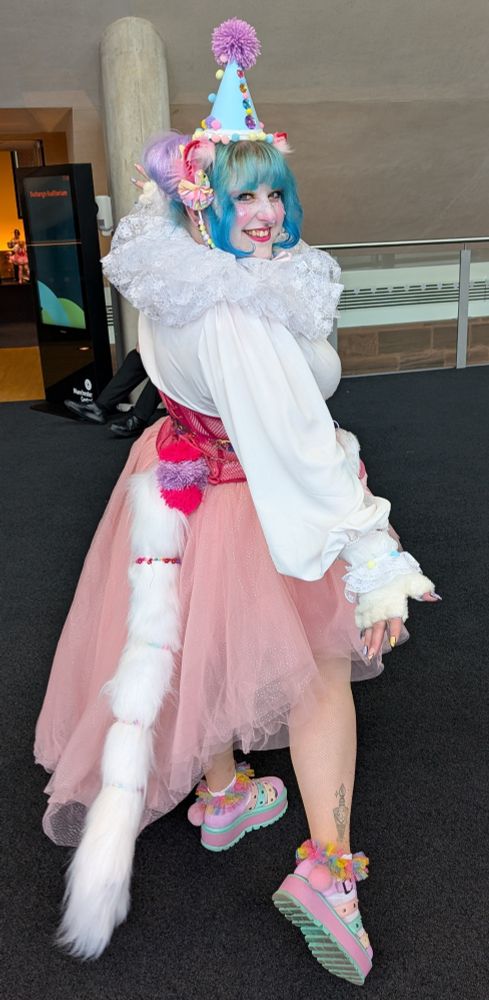 Back view of myself in a blue, purple, pink and yellow party/clown hat, white blouse, pink cat ears, pink corset and skirt and a long white cat tail. 