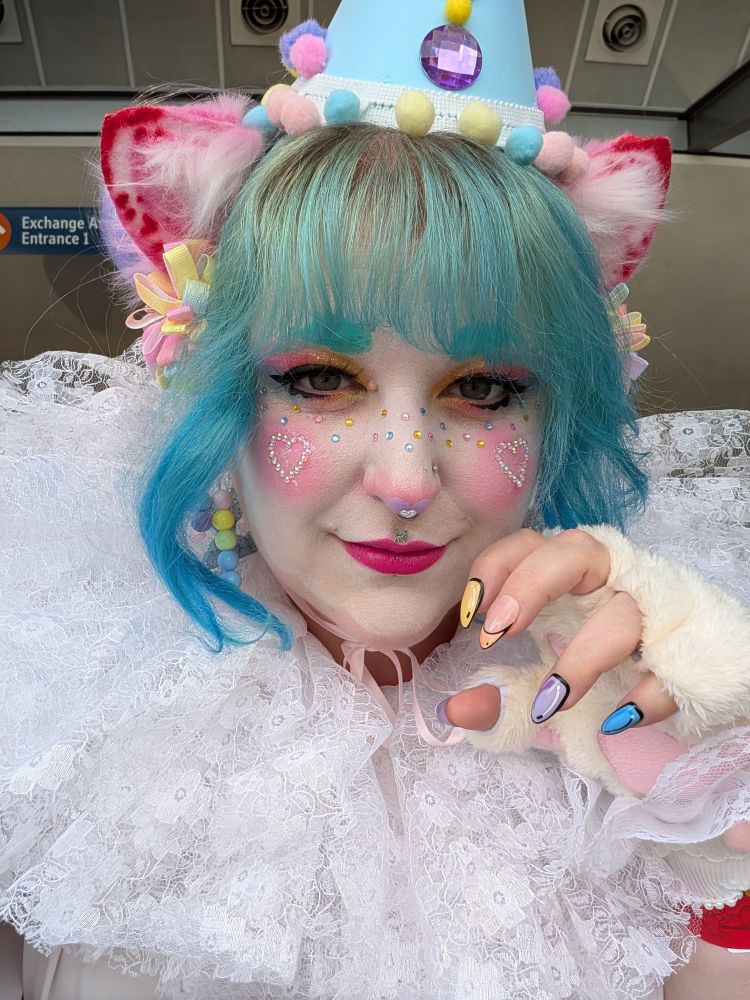 Close up of my clown cat make up, white face paint, pink cheeks with rhinestone hearts on, blue, pink and yellow gem stone freckles, pink lips and yellow, pink and blue eyeshadow. A hand is raised in a claw position showing 2D effect nails 