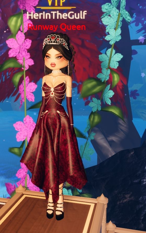The dress to impress model is standing on the podium, in 1st place. She has black, curled hair in a half-up half-down style. She is wearing a patterned, deep red, calf-length dress. The dress has jewels and beaded details draped across the torso. She has strappy black heels on, and a silver tiara on her head. She is also wearing long gloves which go past her elbows, which is the same red pattern as the dress.