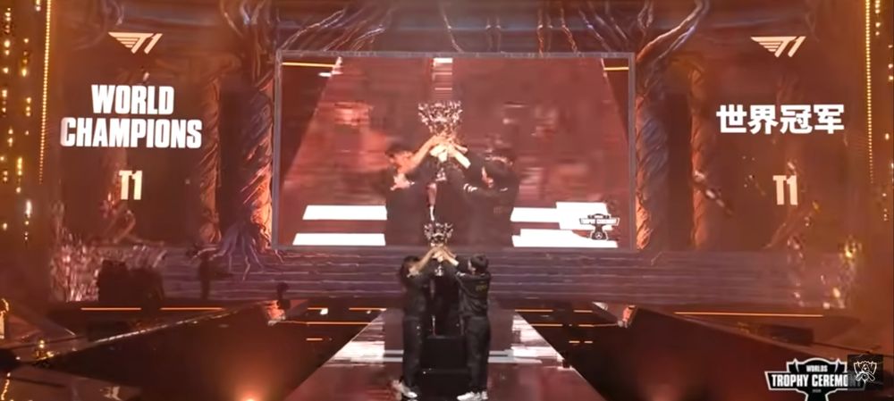 T1 lifting the Summoner Cup above while their name is shown in the background next to World Champions. 