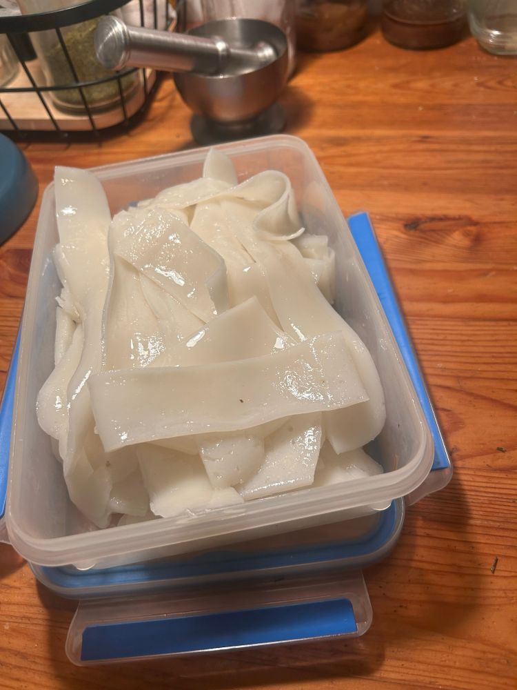 The rice-noodle sheets have been hand cut into narrower strips, lightly oiled so they shine in the light of the table lamp, and heaped into a rectangular Tupperware container. 