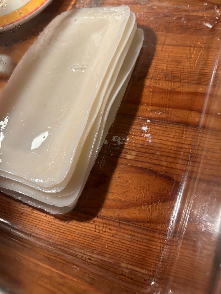 A stack of wide, glossy Rice-noodle sheets in a large, clear glass baking dish, on top of a wooden table.