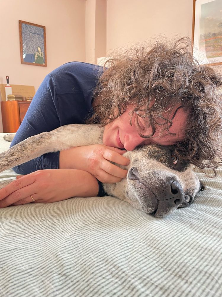 A white Jewish lesbian with curly brown hair falling into her eyes cuddles an adorable blue heeler mix with a brown spot over his one eye and a white and spotted brindle coat.