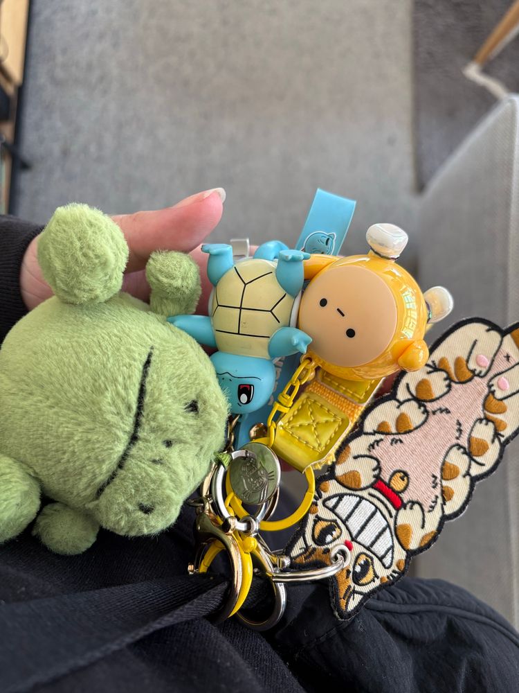 Photo of four keyrings, from left to right: a round stuffed frog with a grumpy face, a vinyl squirtle, a yellow vinyl circle with a face that kind of looks like the teletubby LaLa, and an embroidered catbus from My Neighbour Totoro