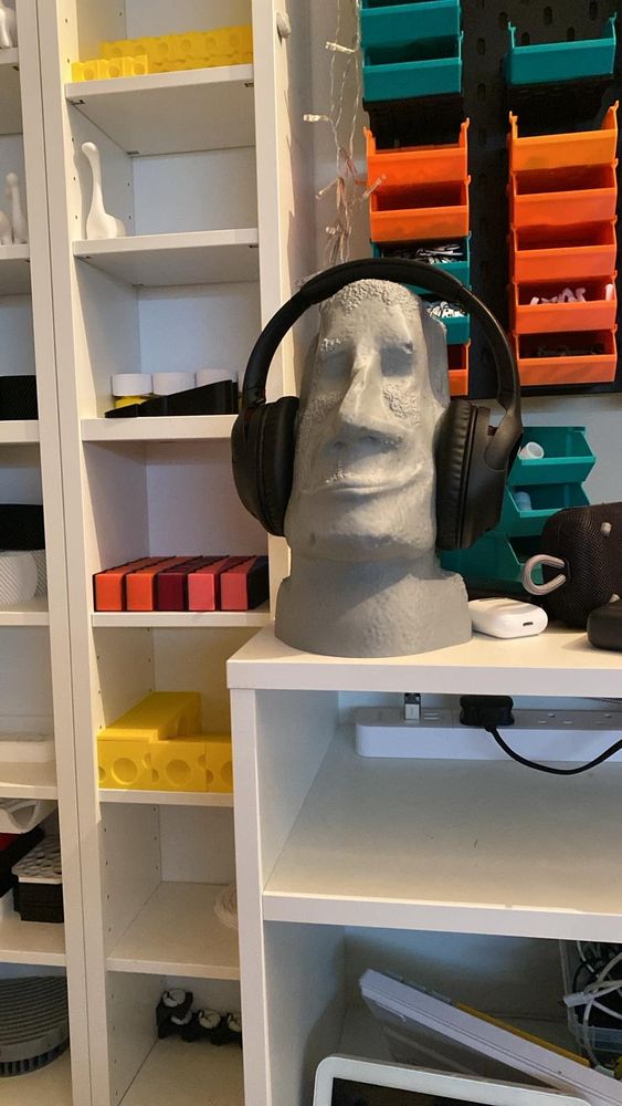 Grey Easter Island bust, with black earphones. Sitting on a white bookcase, surrounded by various colourful shelves