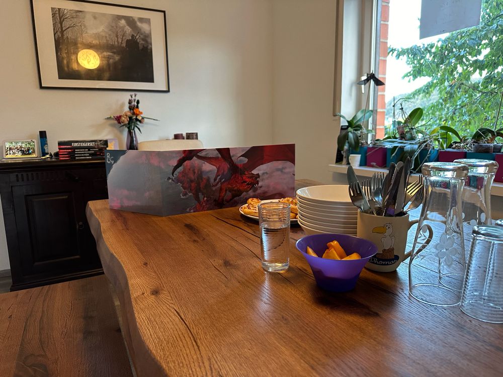 Ein Holztisch. Am Kopfende ein Dungeons & Dragons Meisterschirm. Im Vordergrund Geschirr, Besteck, Gläser und Häppchen. Im Hintergrund ein Bild und ein Fenster ins Grüne
