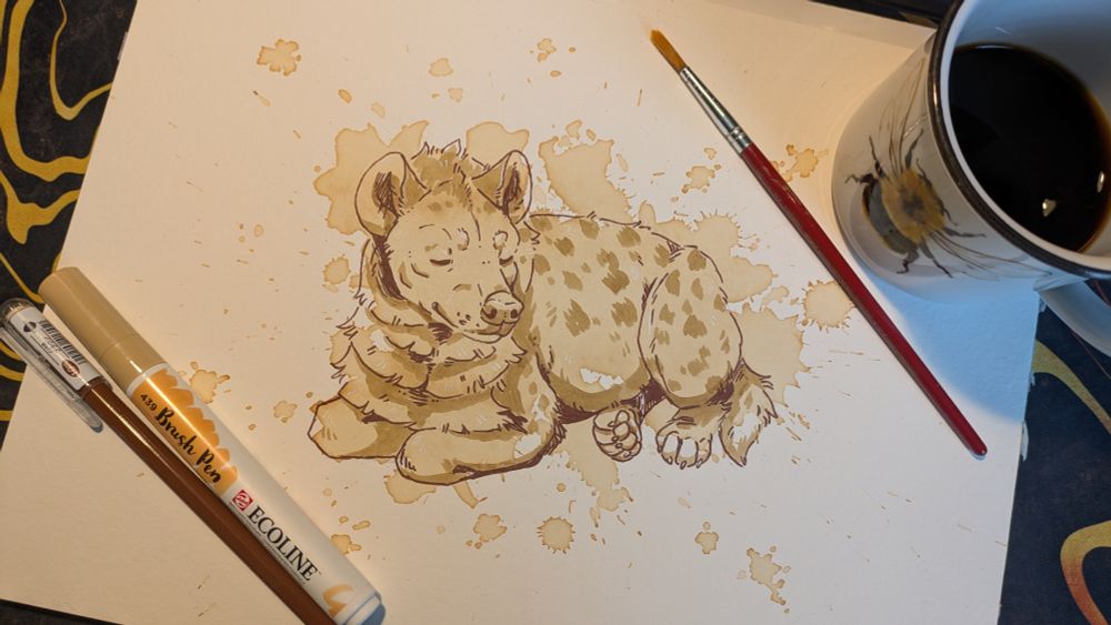 A photo of a piece of paper on a desk. There are a cup of black coffee, a brush, a brown gel pen and a creme brush pen lying on its corners. In the middle is a drawing of a spotted hyena, lying like a loaf with their feet tucked beneath them and closed eyes. The main colour are big, light splotches of dried coffee which have been drawn on with the brown gel pen and creme brush pen to get some shading and the shapes/ lines of the hyena.