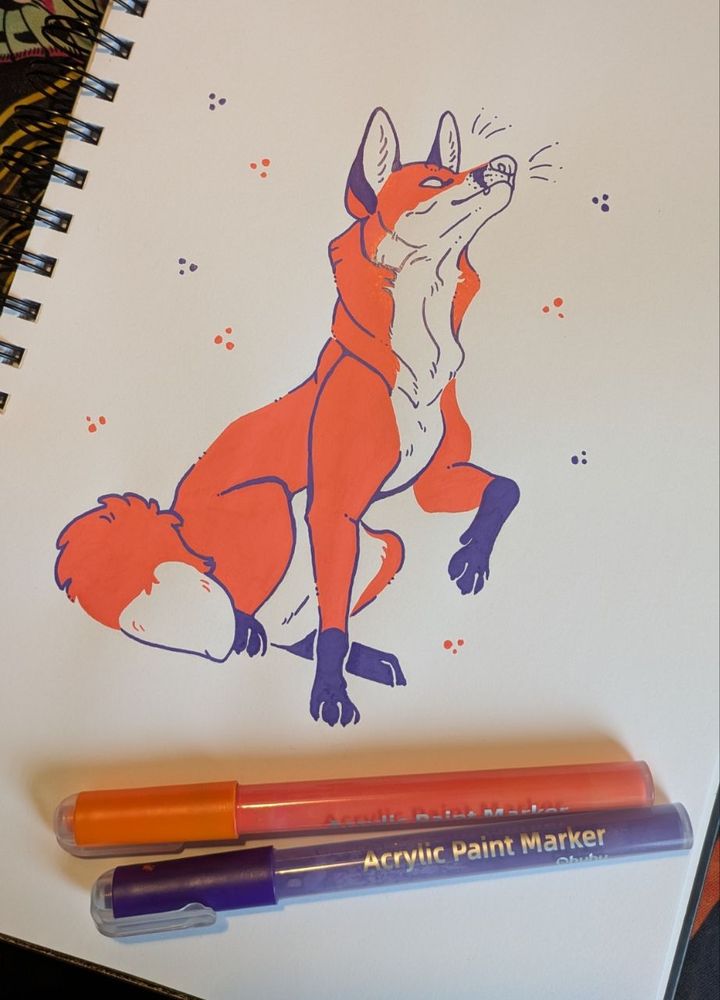 A photo of a sketchbook with 2 acrylic pens lying next to a limited palette picture of a fox. The colours are a vibrant red-orange and a dark violet. The fox is sitting, lifting one leg and looking up.